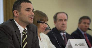 IRS Commissioner Danny Werfel speaks into a microphone while seated at a media podium surrounded by his colleagues.
