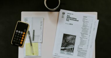An assortment of items rest on a black desk, including official tax withholding forms, a calculator, envelopes, a pen, and cup of coffee.